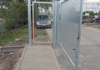 Nowoczesna, ocynkowana brama dwuskrzydłowa z furtką, widok z zewnątrz. Furtka otwarta, w tle zaparkowany samochód. Szara kostka brukowa i fragment ogrodzenia panelowego.