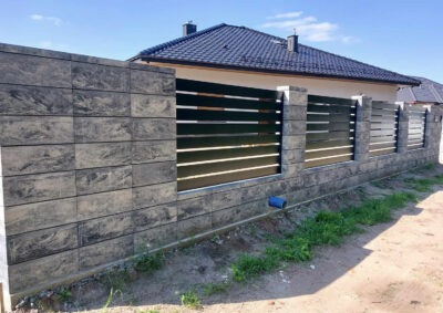 Nowoczesne ogrodzenie panelowe z elementami betonowymi w odcieniach szarości, z widocznym fragmentem domu w tle. Solidna konstrukcja, poziome panele.