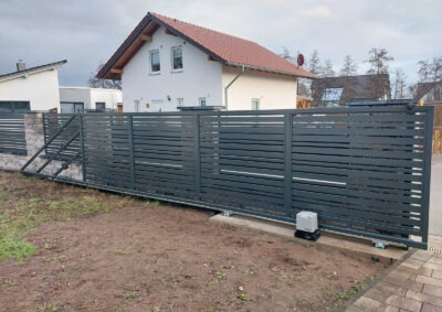 Nowoczesna, przesuwna brama w kolorze antracytowym z automatyką, na tle domu jednorodzinnego. Brama z poziomych profili, widoczny napęd bramy.