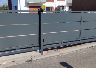 Nowoczesna, przesuwna brama wjazdowa z poziomymi szczelinami, pomalowana na szaro. Widoczny mechanizm jezdny i lampa ostrzegawcza na słupku.