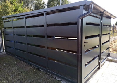 Nowoczesna, grafitowa zabudowa śmietnika z metalowymi panelami i zadaszeniem, z widocznym systemem rynnowym. Estetyczne rozwiązanie na posesji.