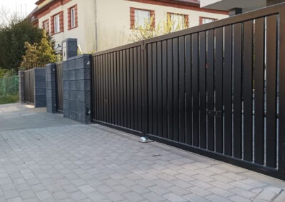 Nowoczesna, przesuwna brama metalowa w kolorze czarnym z pionowymi panelami, osadzona na murowanych słupkach z szarej kostki betonowej na tle domu.