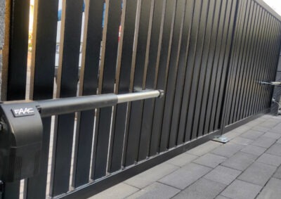 Automatyczna brama wjazdowa z czarnymi, pionowymi panelami i szarą kostką brukową. Widoczny siłownik FAAC. Nowoczesny design.