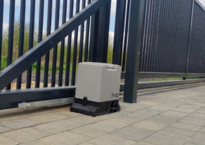 Zamontowany napęd bramy przesuwnej, model 740, przy czarnej, metalowej bramie panelowej na tle kostki brukowej i zieleni.