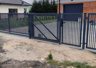 Nowoczesna, grafitowa brama przesuwna i furtka z pionowych profili na tle domu z garażem. Widoczne mocowania i zamek furtki. Minimalistyczny design.