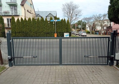 Nowoczesna, metalowa brama wjazdowa z automatyką, szara, pionowe panele. W tle budynki mieszkalne i zieleń. Brama dwuskrzydłowa na posesji.