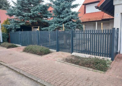Grafitowe ogrodzenie panelowe z furtką, widok z chodnika na tle domu z czerwonym dachem i zielonymi drzewami. Wzdłuż ogrodzenia rosną krzewy.