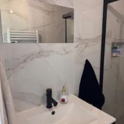 Umywalka w nowoczesnej łazience z marmurowymi płytkami, czarną armaturą i akcesoriami. Widoczny fragment kabiny prysznicowej i ręcznik.