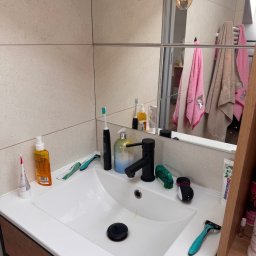 Vina grup - Umywalka z czarną armaturą, otoczona kosmetykami i akcesoriami higienicznymi. W tle lustro odbijające ręczniki i lampy. Widok na fragment łazienki.