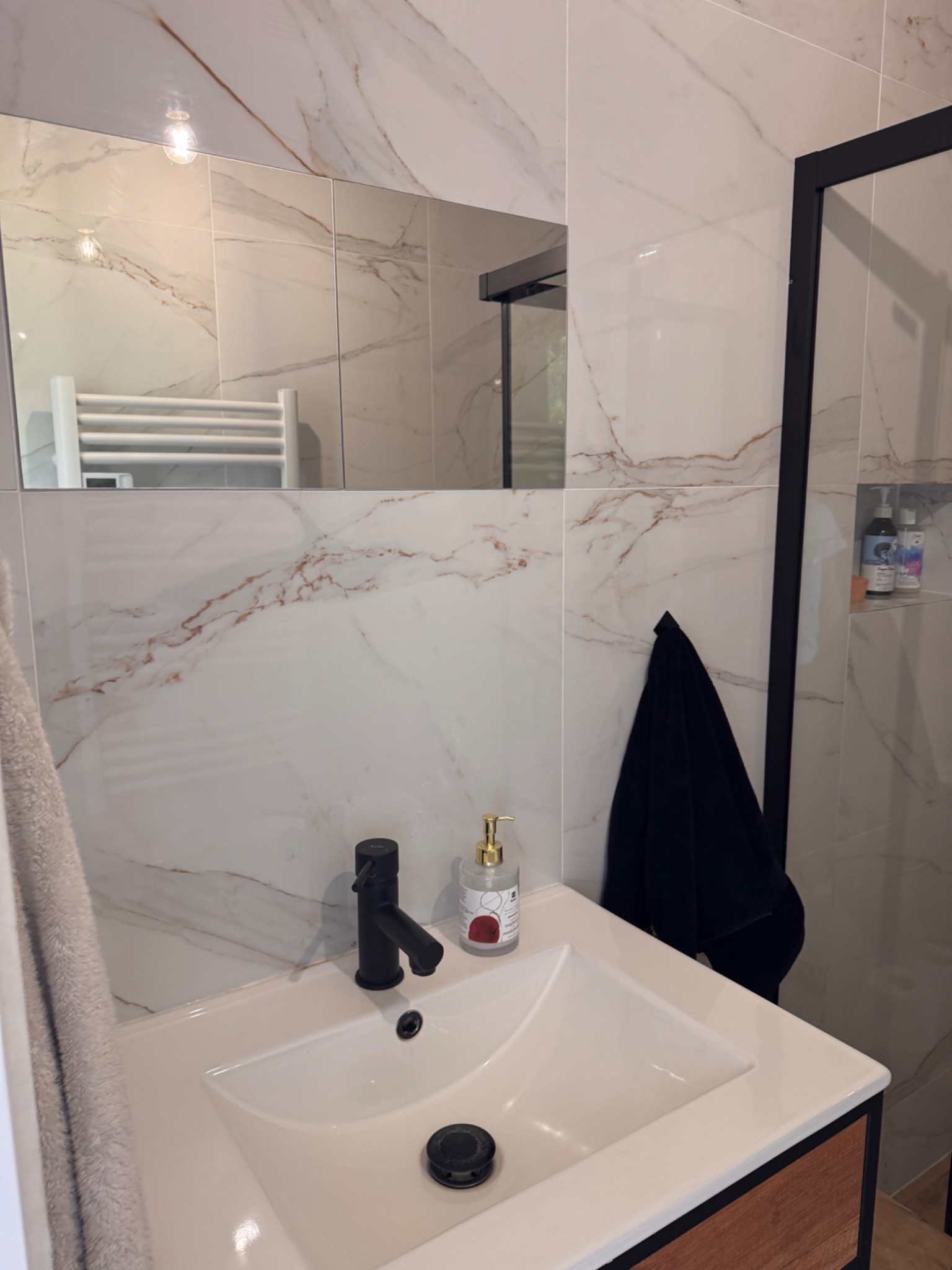 Umywalka w nowoczesnej łazience z marmurowymi płytkami, czarną armaturą i akcesoriami. Widoczny fragment kabiny prysznicowej i ręcznik.