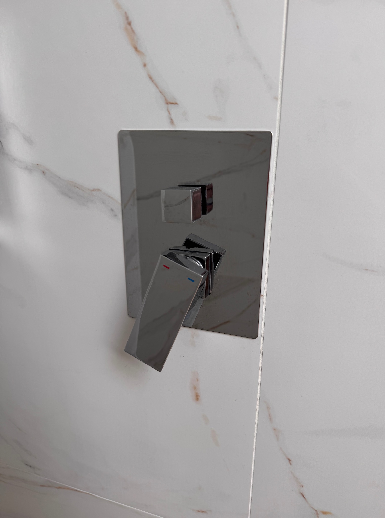 Bateria prysznicowa w chromowanym wykończeniu, zamontowana na białej marmurowej ścianie z subtelnymi brązowymi żyłkami. Minimalistyczny design.