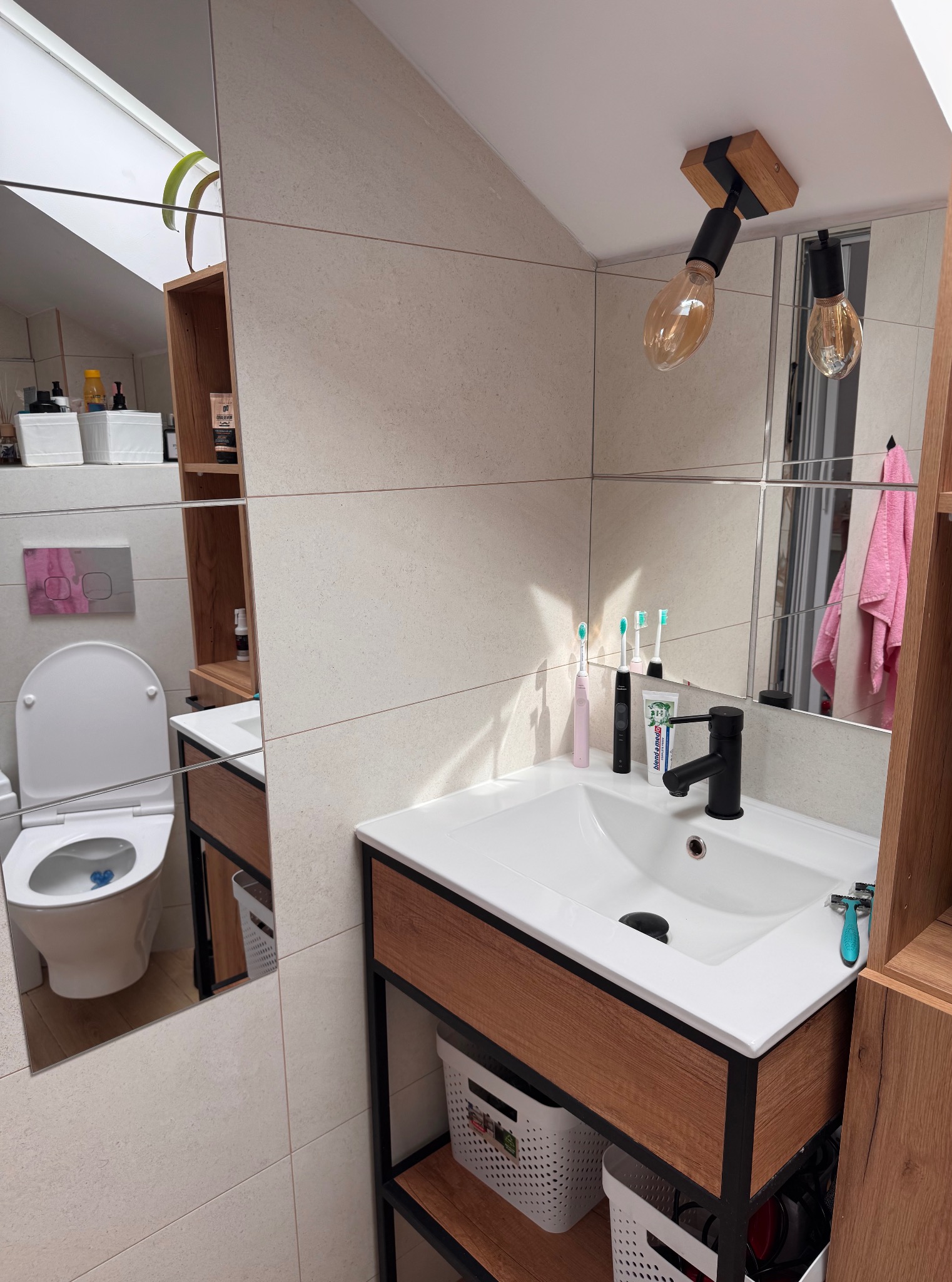 Nowoczesna łazienka na poddaszu z umywalką, toaletą i drewnianymi akcentami. Nietypowe oświetlenie w postaci żarówek. Ujęcie z perspektywy pod kątem.