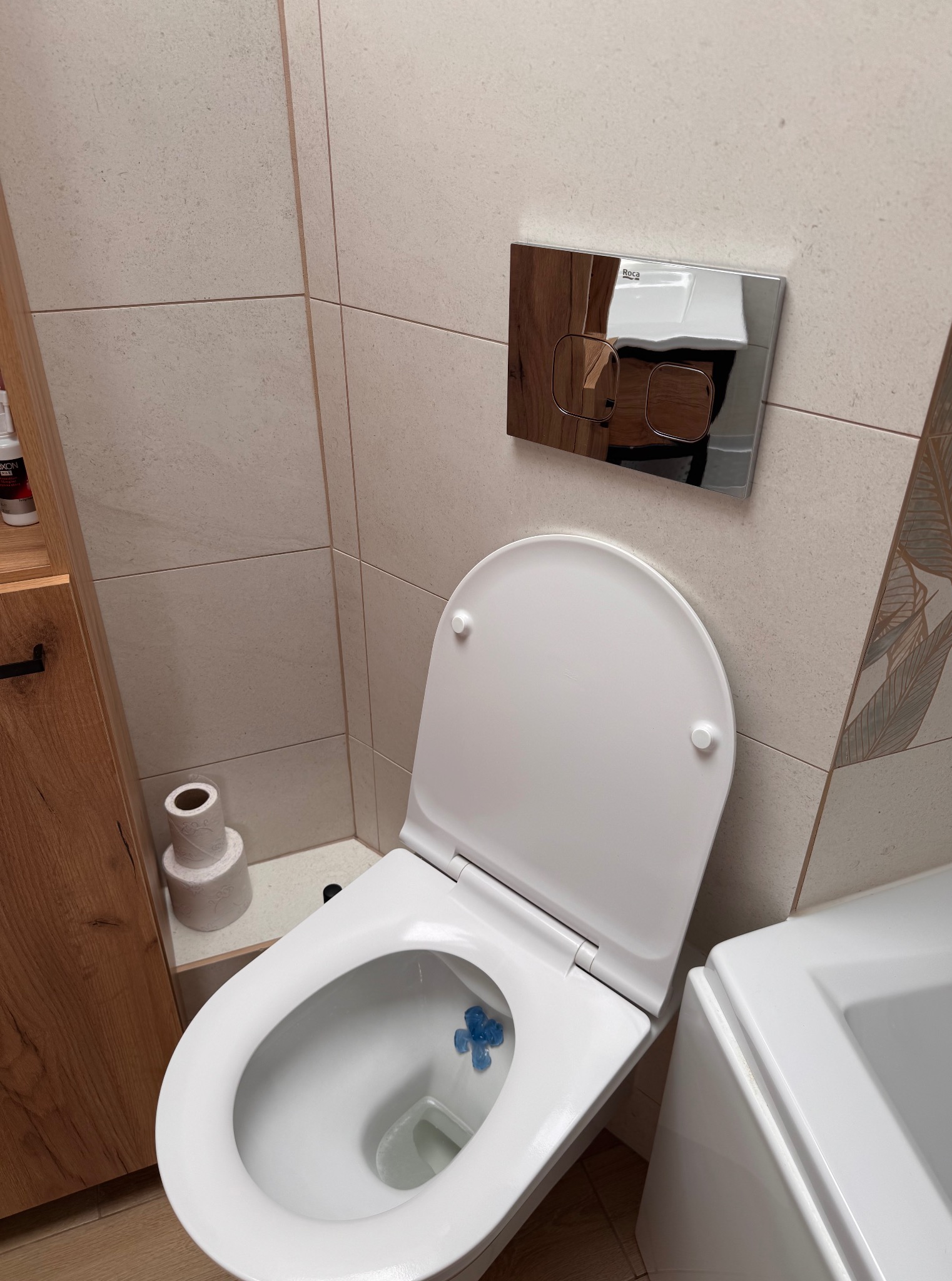 Biała toaleta z odświeżaczem w nowoczesnej łazience z beżowymi płytkami i drewnianą szafką. Widoczny dozownik papieru toaletowego i chromowany przycisk spłukiwania.