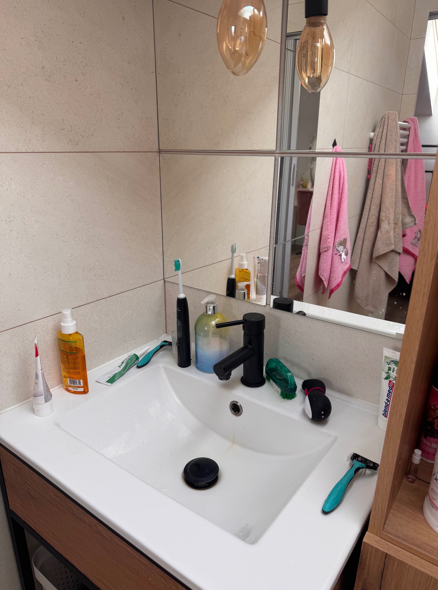 Umywalka z czarną armaturą, otoczona kosmetykami i akcesoriami higienicznymi. W tle lustro odbijające ręczniki i lampy. Widok na fragment łazienki.