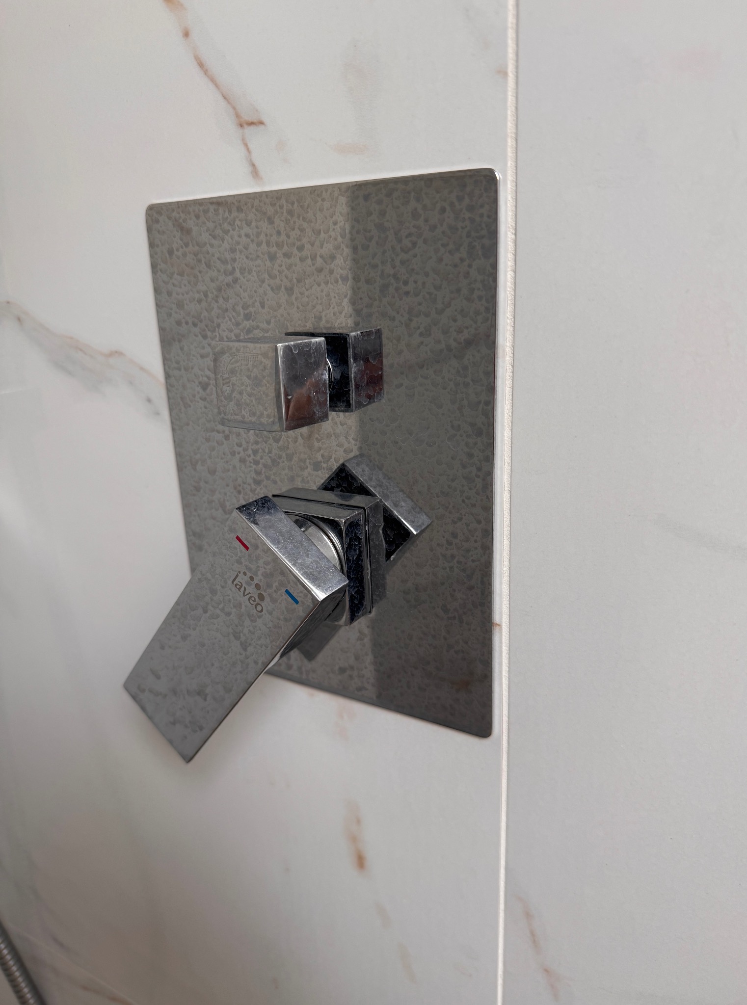 Bateria prysznicowa Laveo z kwadratową rozetą na białej płytce imitującej marmur z delikatnym, beżowym żyłowaniem. Widoczne krople wody.