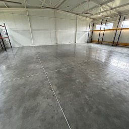 Radosław Zieliński Posadzki - Błyszcząca, szara posadzka w hali magazynowej, z widocznymi liniami podziału. Metalowa konstrukcja dachu i regały w tle. Minimalistyczny, industrialny wygląd.