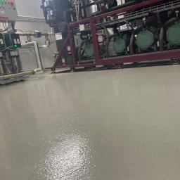 Radosław Zieliński Posadzki - Błyszcząca, szara powierzchnia podłogi w hali przemysłowej, z widocznymi elementami instalacji i urządzeń chłodniczych w tle. Gładka, jednolita struktura posadzki.