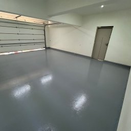 Radosław Zieliński Posadzki - Gładka, szara posadzka w garażu z otwartą bramą segmentową i drzwiami wejściowymi. Błyszcząca powierzchnia, minimalistyczne wnętrze, neutralna kolorystyka.