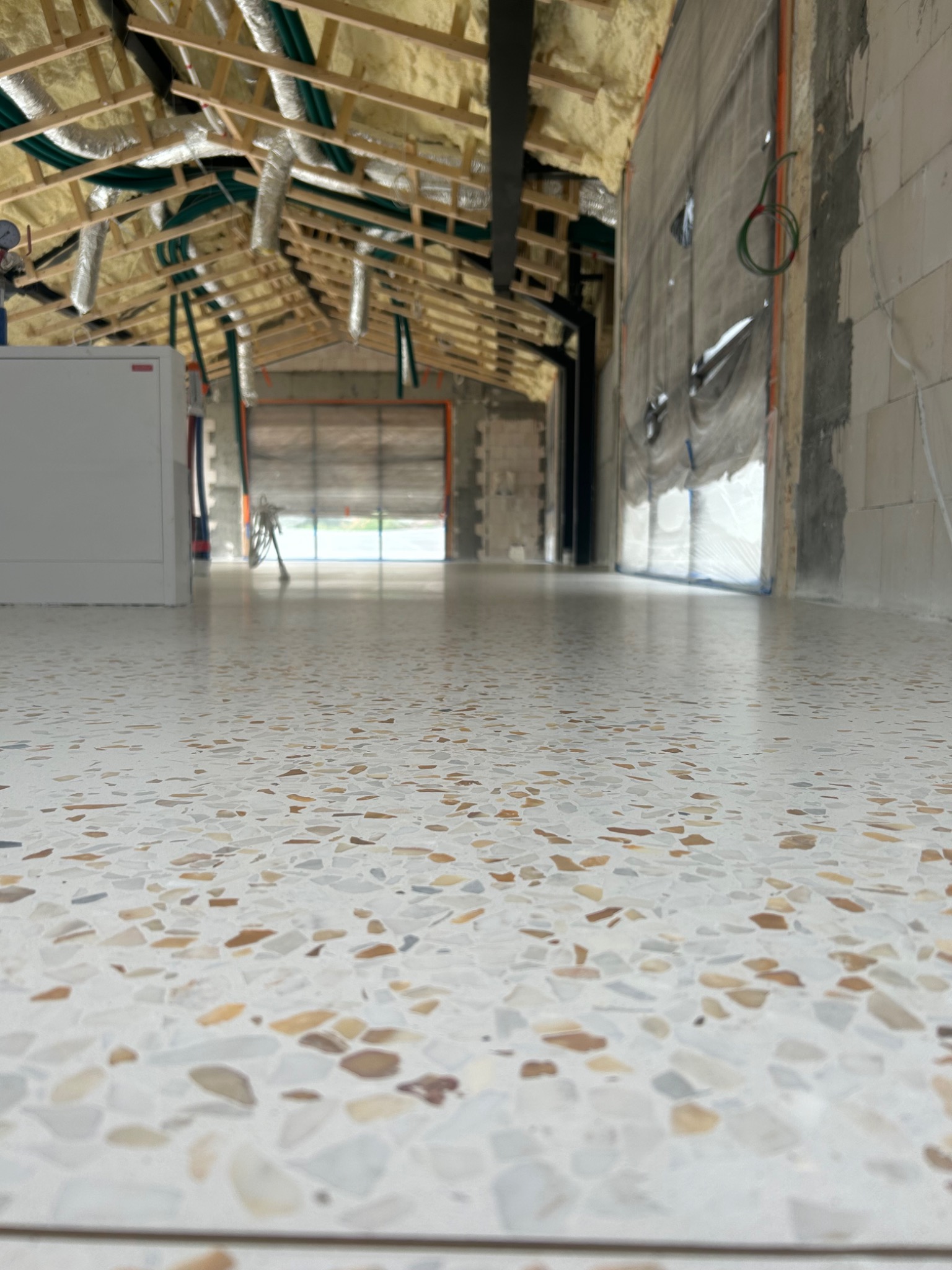 Biała posadzka z kamiennym wzorem w surowym wnętrzu hali. Widoczne instalacje pod stropem i ściany w trakcie wykończenia. Perspektywa z niskiego kąta podkreśla gładką powierzchnię.