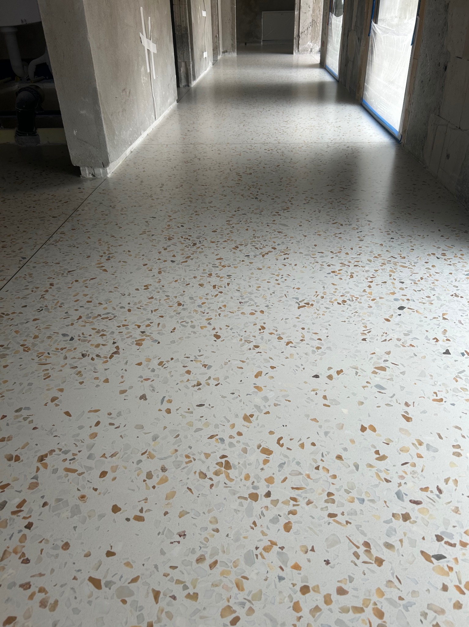 Jasna posadzka lastryko z drobnymi, kolorowymi kamyczkami w korytarzu budynku w stanie deweloperskim. Widoczne surowe ściany i przygotowania do wykończenia.