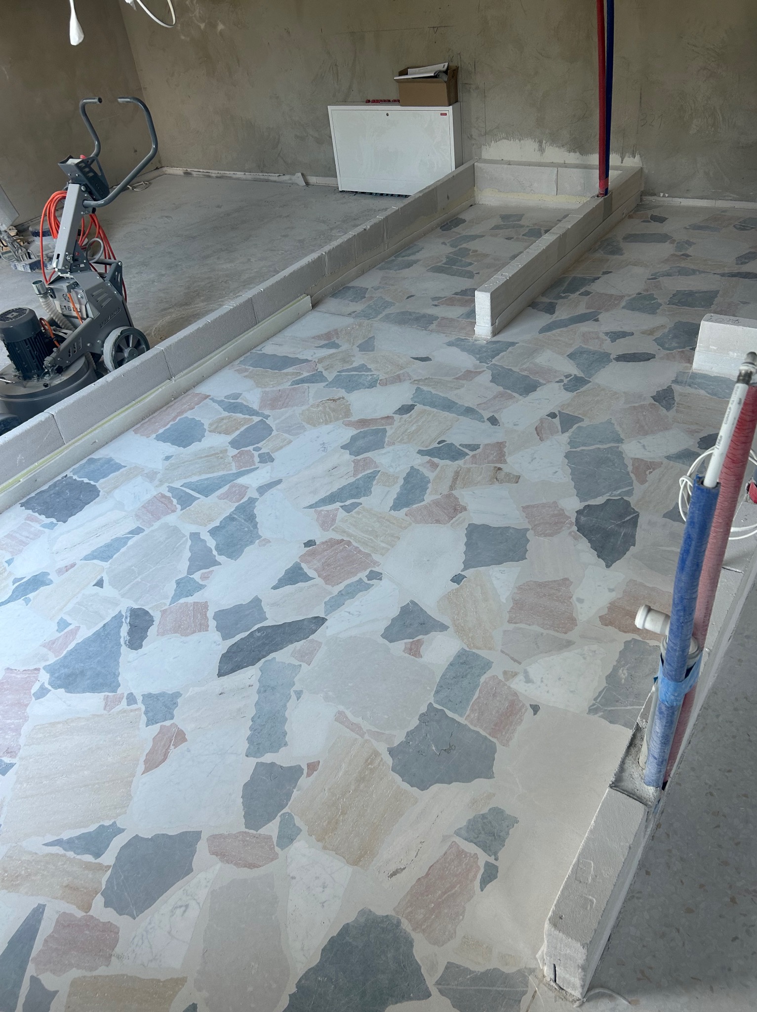 Polerowana posadzka lastryko z nieregularnymi wzorami, obramowana szarymi krawężnikami. W tle maszyna do polerowania i elementy instalacji hydraulicznej.