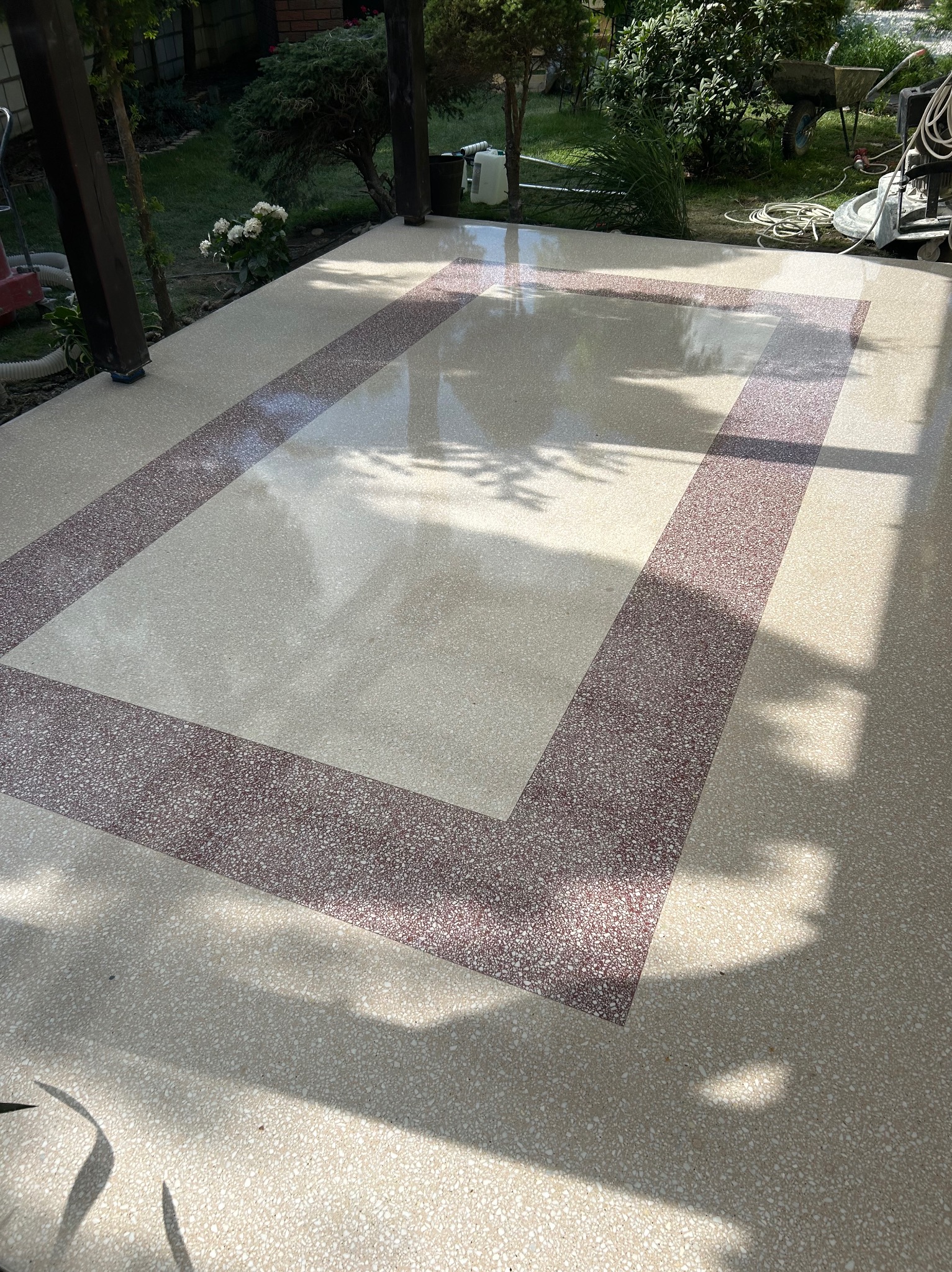 Błyszcząca, beżowa posadzka żywiczna z bordową obwódką na zewnątrz, na tarasie. Widoczne narzędzia pracy i cień drzewa. Nowoczesny design.