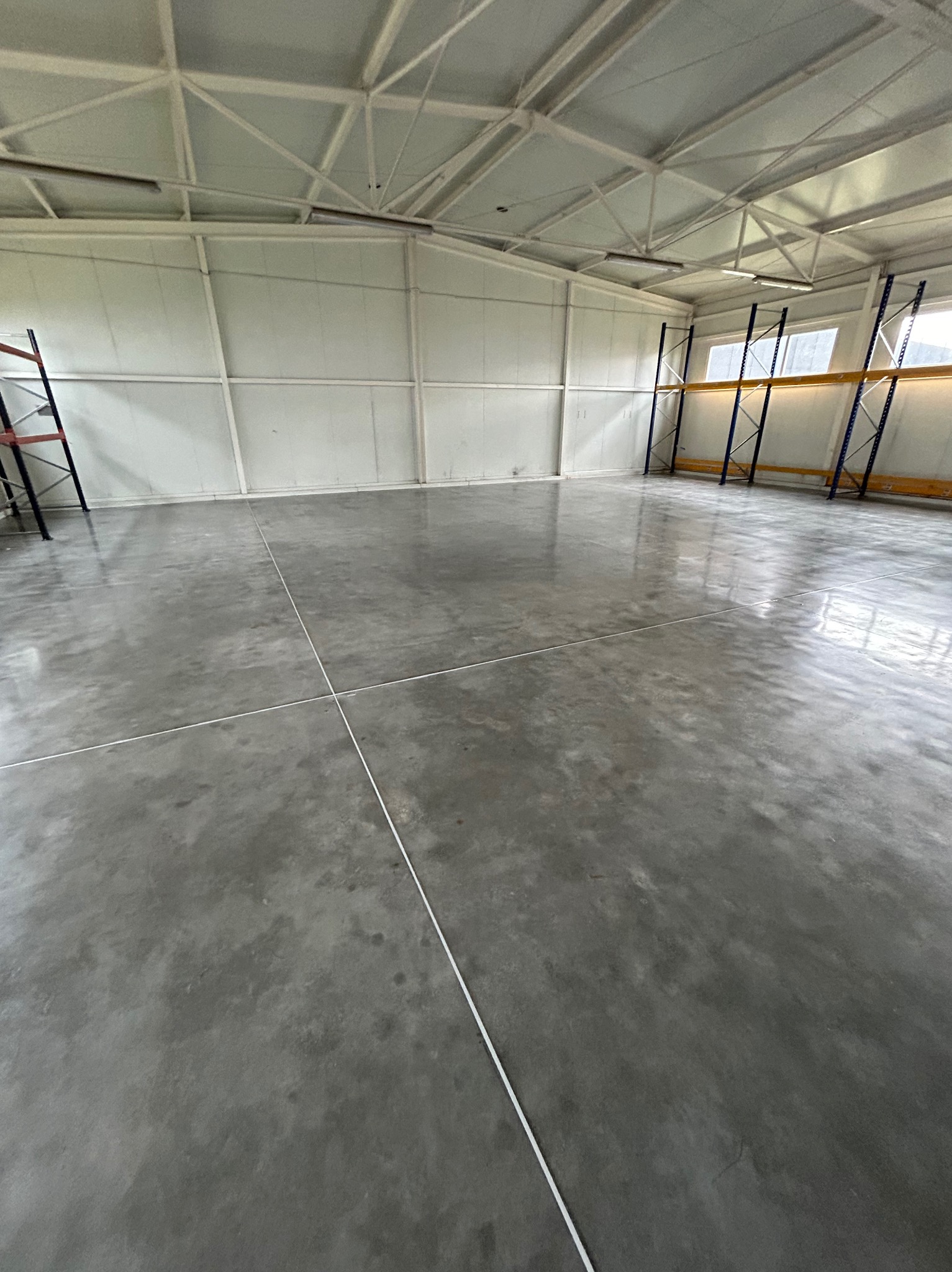 Błyszcząca, szara posadzka w hali magazynowej, z widocznymi liniami podziału. Metalowa konstrukcja dachu i regały w tle. Minimalistyczny, industrialny wygląd.