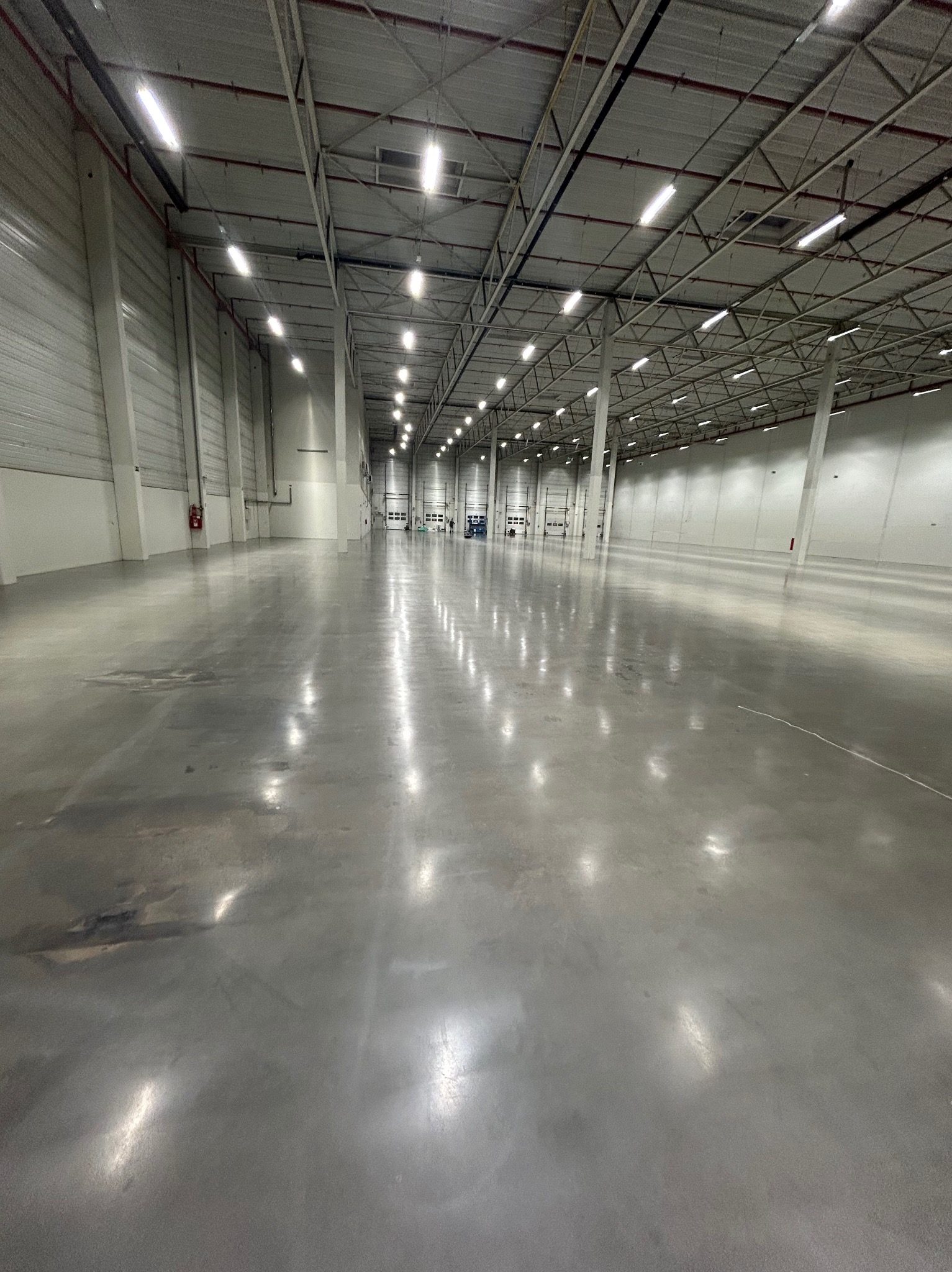 Błyszcząca, szara posadzka w przestronnej hali magazynowej z białymi ścianami i rzędem lamp sufitowych. Widoczne ślady po procesie obróbki.