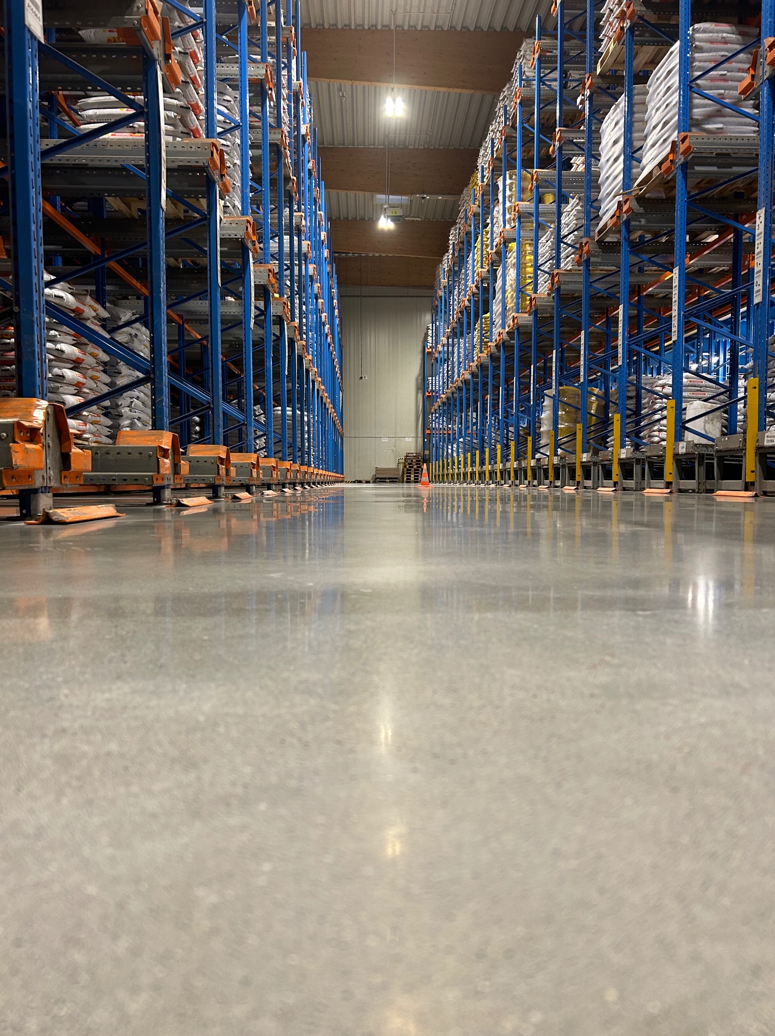 Błyszcząca, szara posadzka w magazynie z wysokimi regałami w kolorze niebieskim i pomarańczowym, wypełnionymi towarem. Widok z niskiej perspektywy.