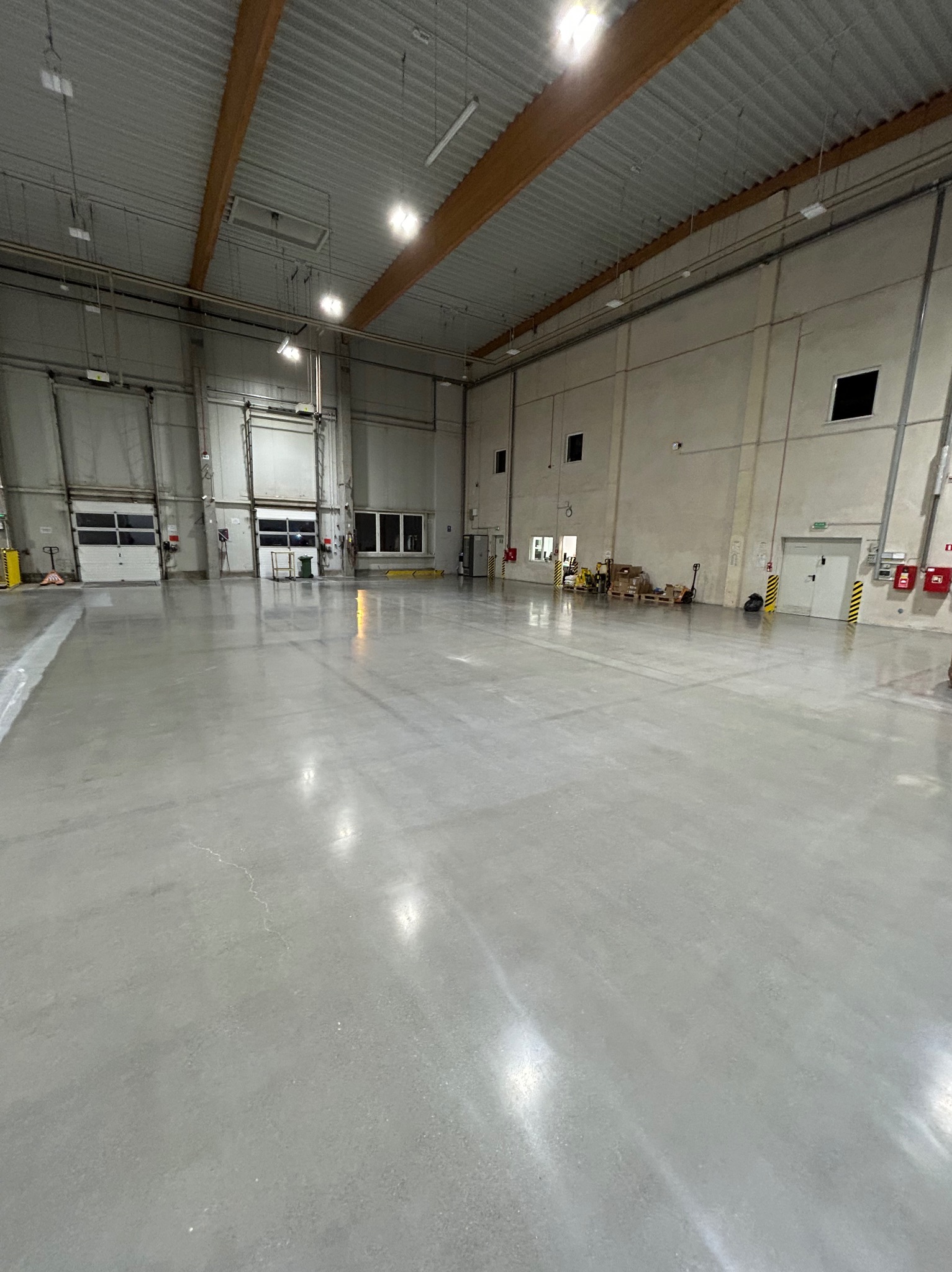 Błyszcząca, szara posadzka w przestronnym magazynie z bramami i oknami. Widoczne żółte zabezpieczenia i sprzęt w tle, podkreślające przemysłowy charakter.