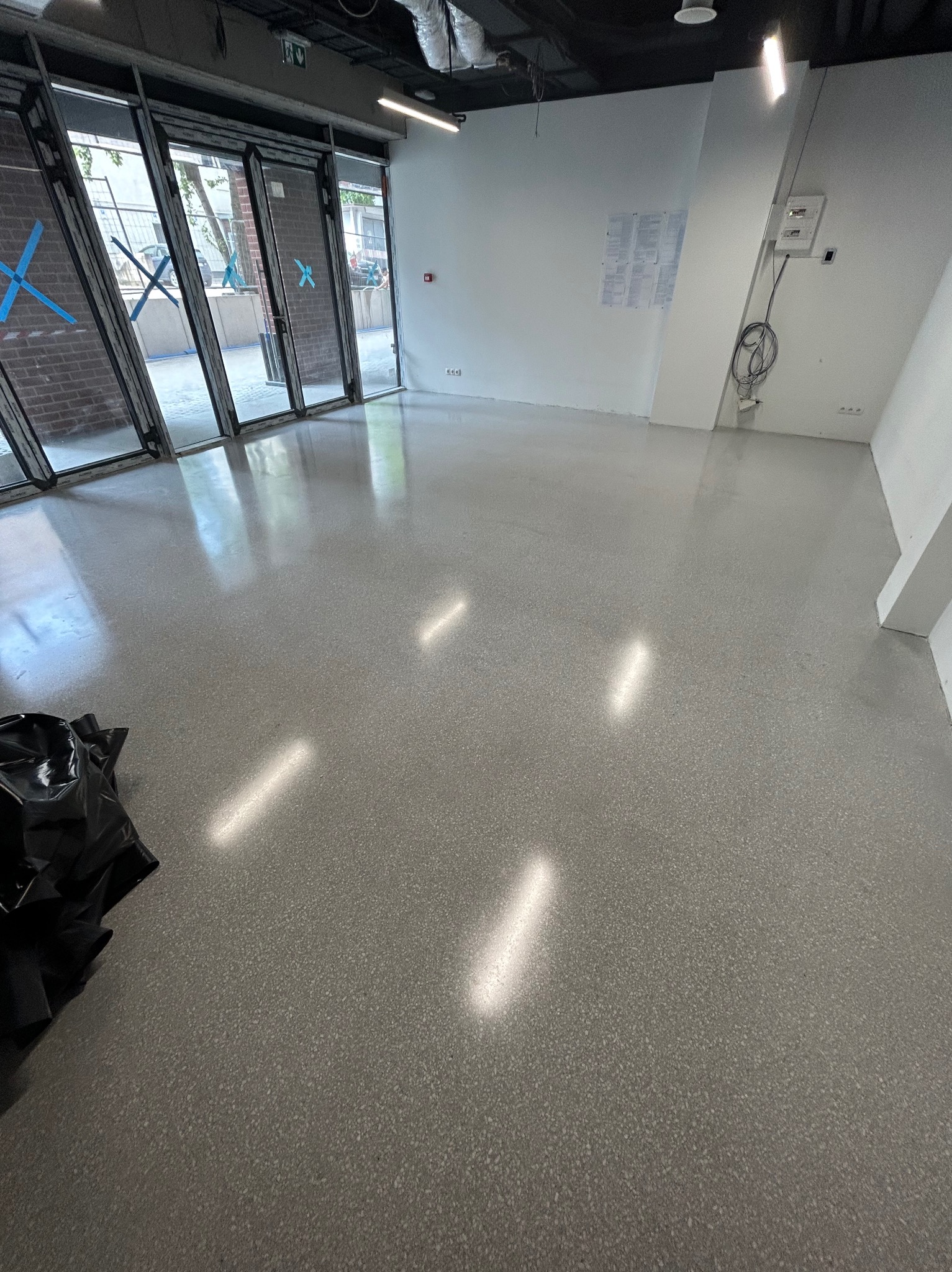 Błyszcząca, szara posadzka w jasnym pomieszczeniu z dużymi oknami. Widoczne refleksy świetlne i worek z odpadami w rogu. Minimalistyczne wnętrze.