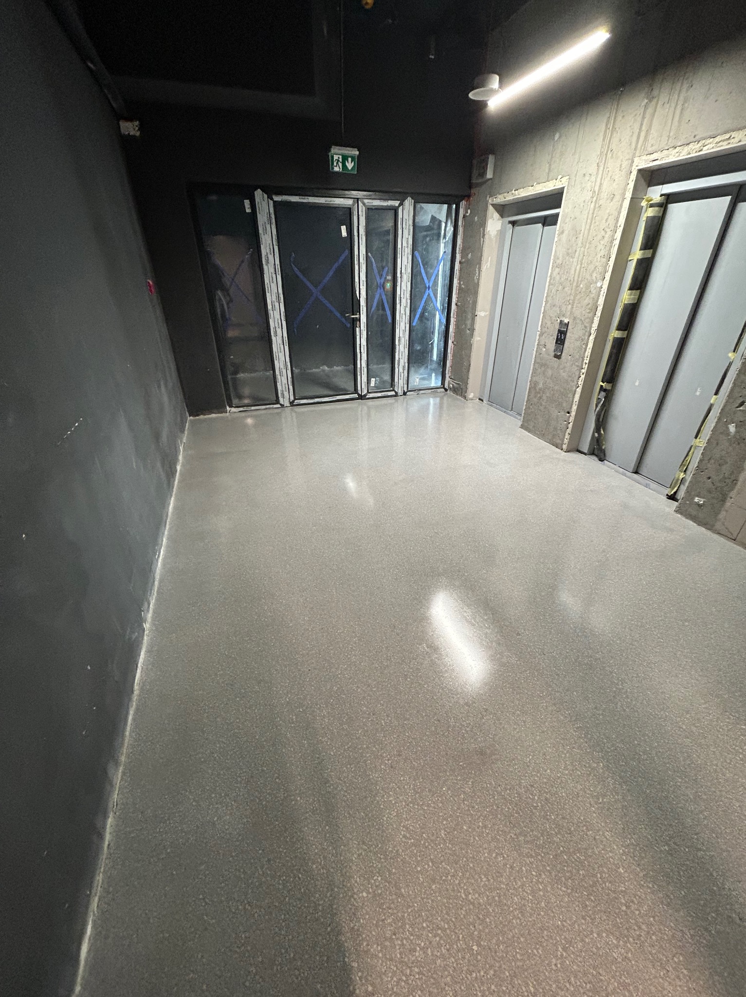 Szara, gładka posadzka przemysłowa w surowym wnętrzu budynku z windami i szklanymi drzwiami zabezpieczonymi taśmą.