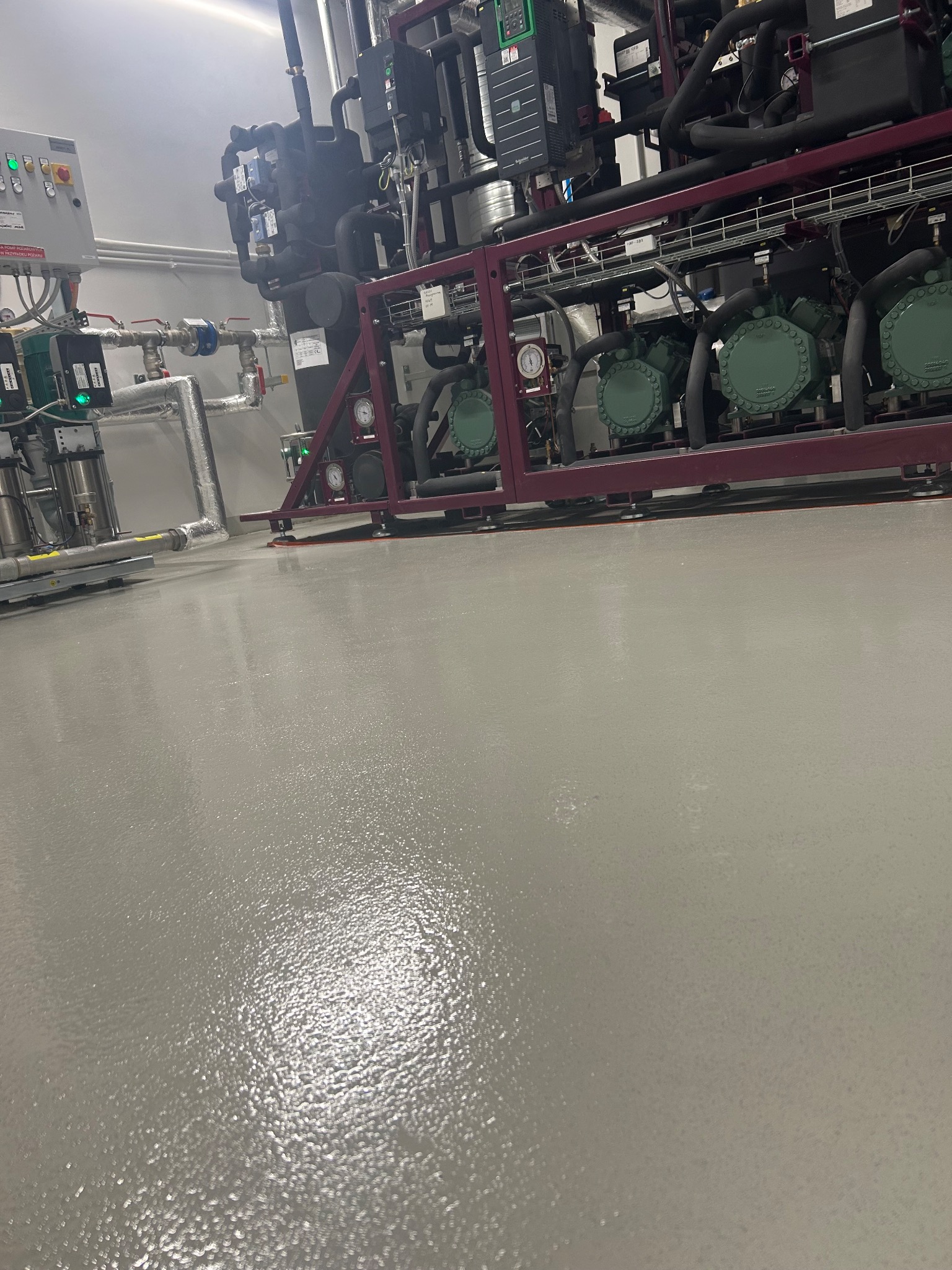 Błyszcząca, szara powierzchnia podłogi w hali przemysłowej, z widocznymi elementami instalacji i urządzeń chłodniczych w tle. Gładka, jednolita struktura posadzki.