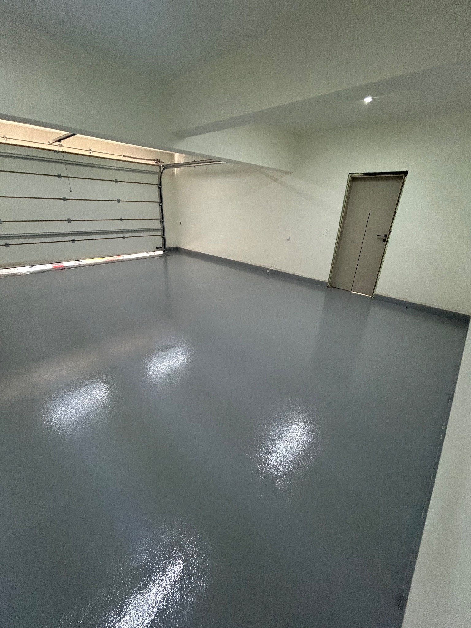Gładka, szara posadzka w garażu z otwartą bramą segmentową i drzwiami wejściowymi. Błyszcząca powierzchnia, minimalistyczne wnętrze, neutralna kolorystyka.
