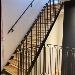 smk.pl Krzysztof Smyczyński - Nowoczesne schody metalowe z drewnianymi stopniami, czarne poręcze i balustrada z pionowymi elementami, jasne ściany i podłoga z parkietem w jodełkę.