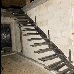 smk.pl Krzysztof Smyczyński - Surowe, metalowe schody z profili kwadratowych, w trakcie montażu w surowym wnętrzu z białych bloczków. Widoczne kable elektryczne na podłodze i przygotowane mocowania.