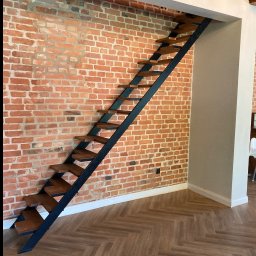 smk.pl Krzysztof Smyczyński - Metalowe schody z drewnianymi stopniami na tle ściany z czerwonej cegły i podłogi w jodełkę. Nowoczesny design, minimalistyczny styl, dobrze oświetlone wnętrze.