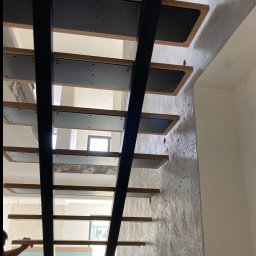 smk.pl Krzysztof Smyczyński - Nowoczesne schody z czarną metalową konstrukcją i drewnianymi stopniami, widok z dołu, podczas montażu, z widocznym fragmentem ściany o dekoracyjnej fakturze.