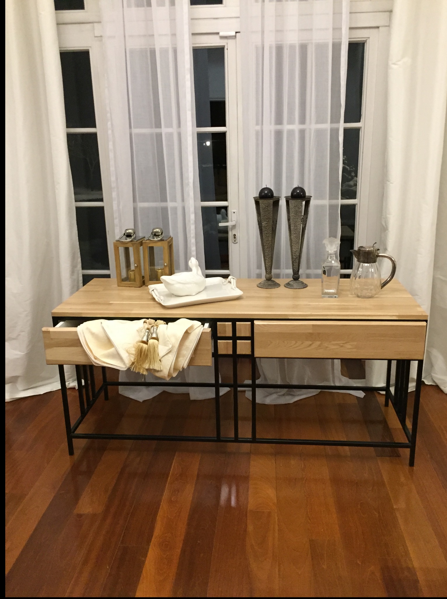 Stół z jasnego drewna i czarną metalową konstrukcją, z wysuwaną szufladą i dekoracjami, na tle okna z białymi zasłonami i drewnianej podłogi.