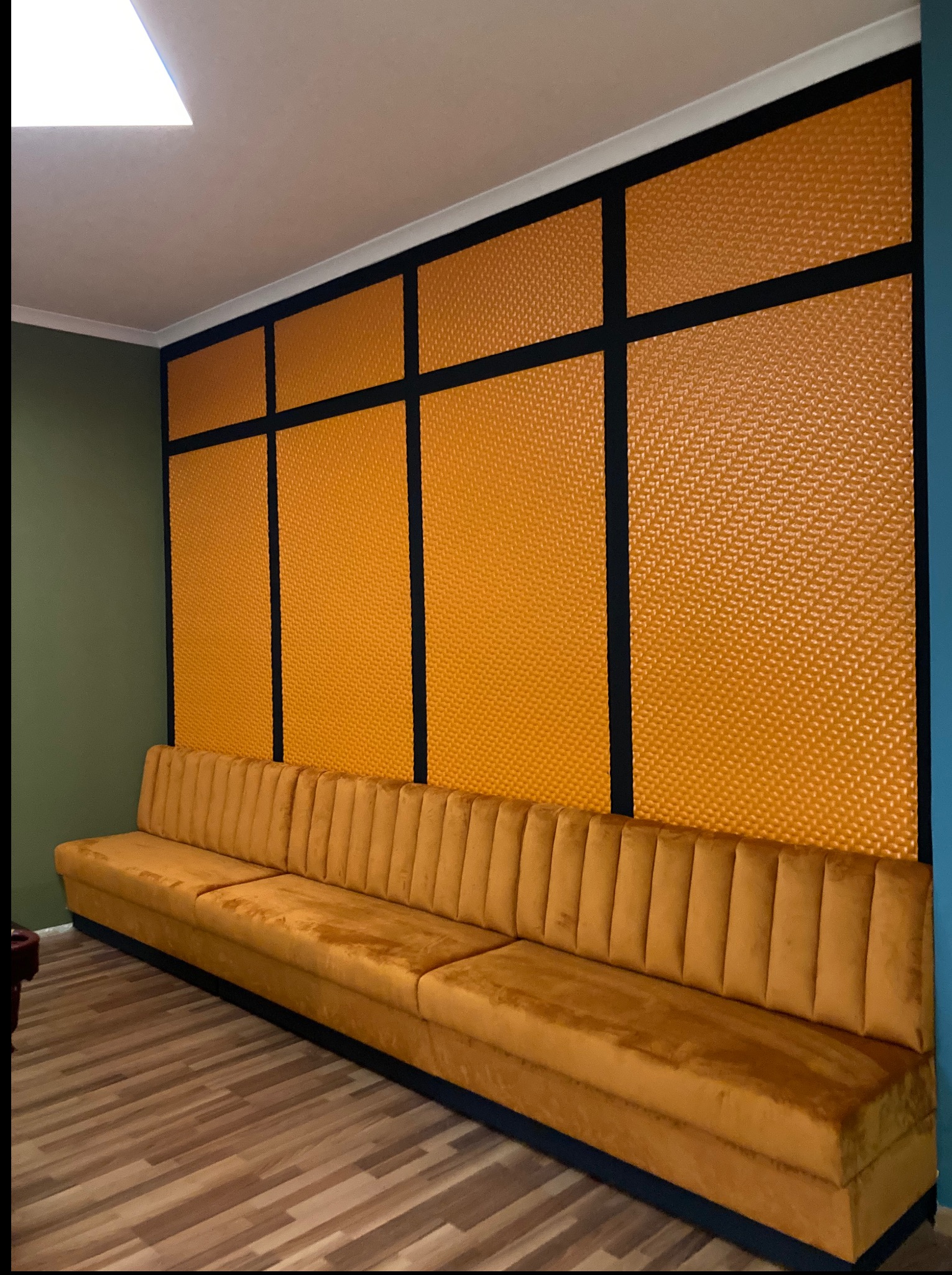 Żółta, pikowana ściana z czarnymi, metalowymi elementami tworzącymi geometryczny wzór, uzupełniona o pasującą, aksamitną kanapę i drewnianą podłogę.