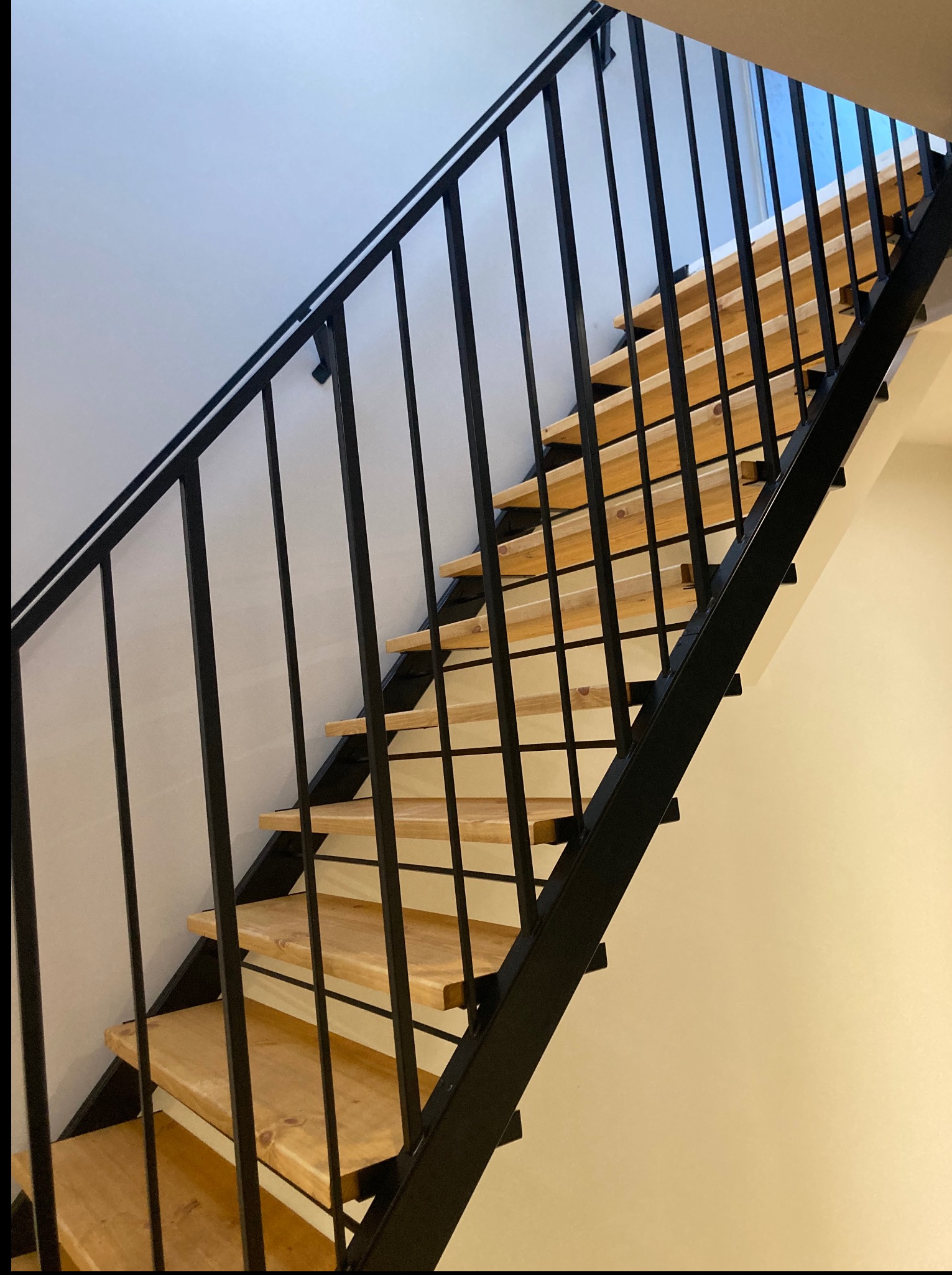 Nowoczesne schody z drewnianymi stopniami i czarną, metalową balustradą z pionowymi prętami, widok z dołu, oświetlenie dzienne.