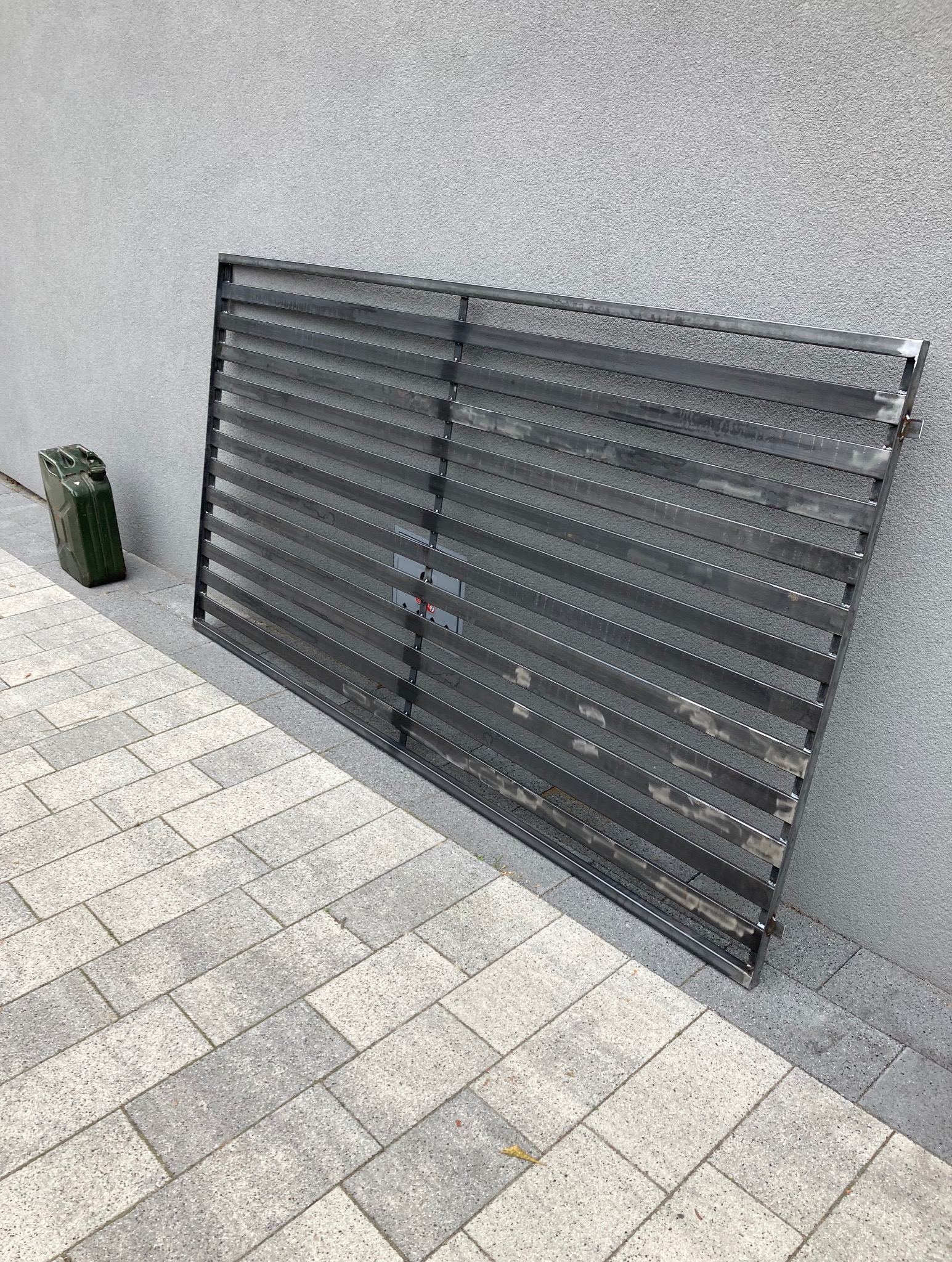 Metalowa, surowa brama z poziomych profili, oparta o szarą ścianę i chodnik z kostki. Obok kanister wojskowy. Widoczne ślady spawania i obróbki.