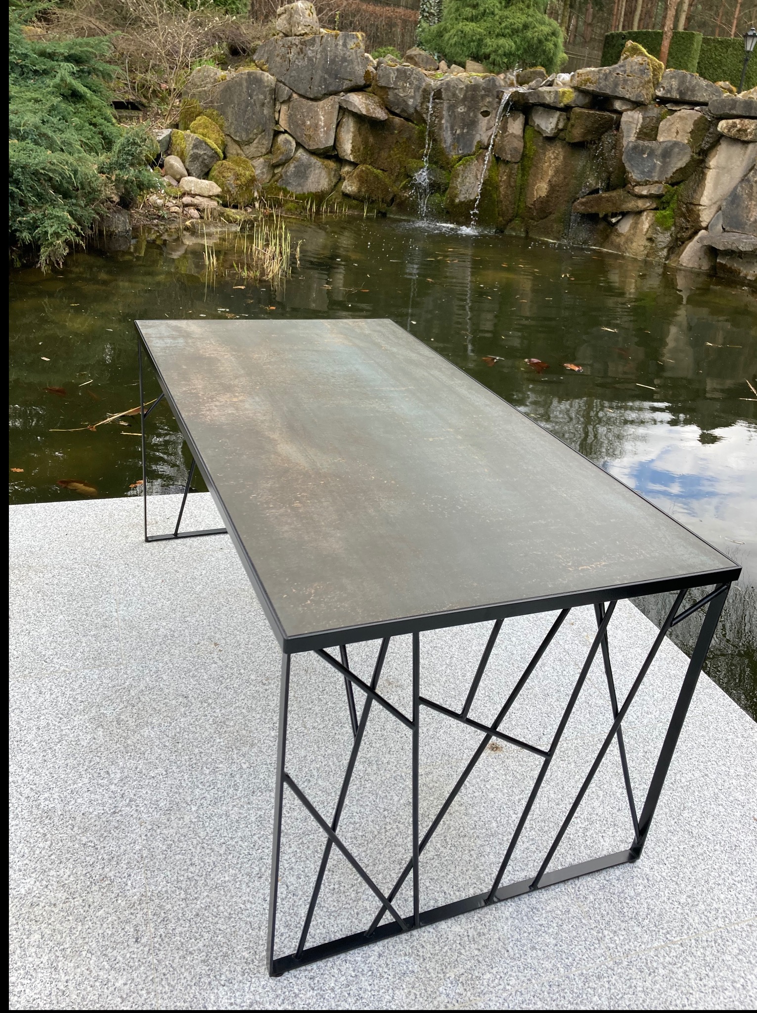 Metalowy stół z geometryczną podstawą na tle kaskady wodnej i zieleni. Blat stołu ma szary, lekko rdzawy odcień, kontrastujący z czarną, ażurową konstrukcją nóg.