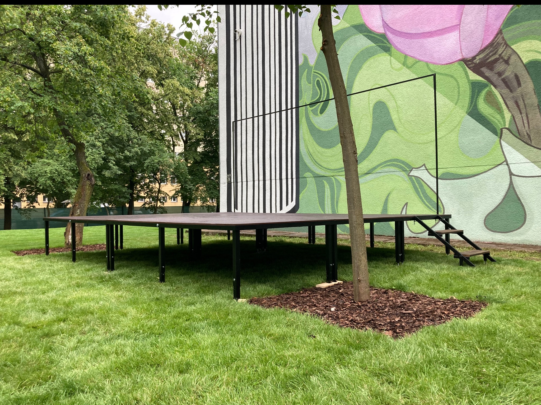 Metalowa konstrukcja podestu/sceny na trawie, z fragmentem schodów i drzewami w tle, przed ścianą z abstrakcyjnym malunkiem. Widok z poziomu gruntu.