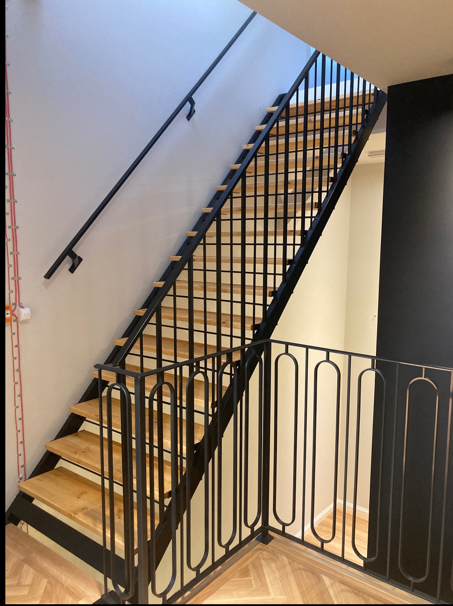 Nowoczesne schody metalowe z drewnianymi stopniami, czarne poręcze i balustrada z pionowymi elementami, jasne ściany i podłoga z parkietem w jodełkę.