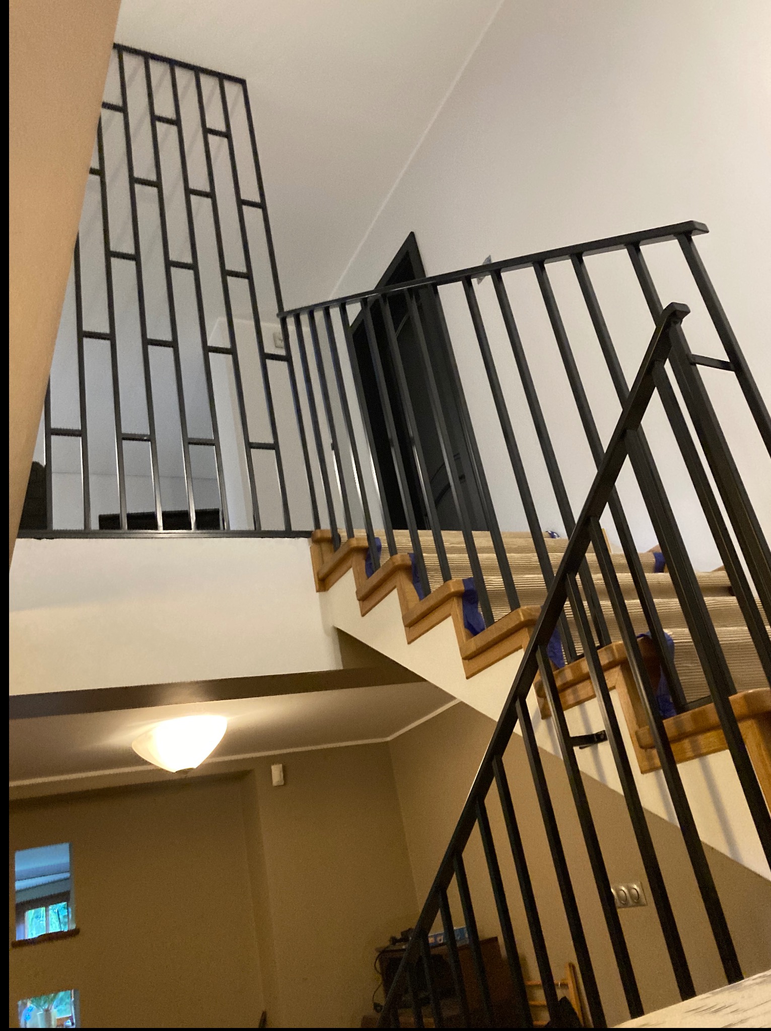 Metalowe schody z czarną balustradą i poręczą w domu w Toruniu. Nowoczesny design z pionowymi szczeblami. Widok z dołu, oświetlenie sufitowe.