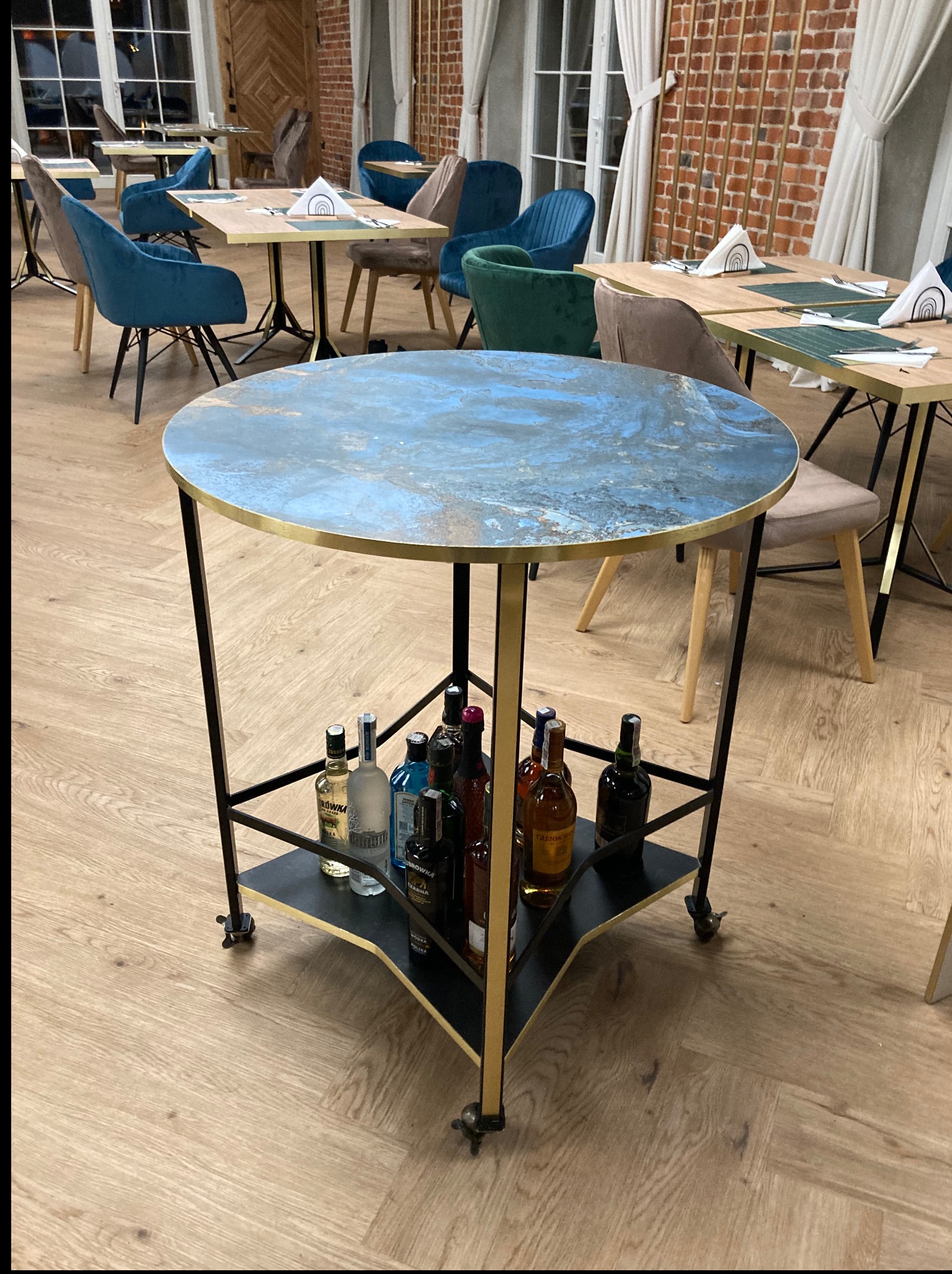 Stylowy, okrągły stolik barowy na kółkach z blatem imitującym marmur, z półką na butelki alkoholu. W tle widoczne inne stoliki i krzesła w restauracji.