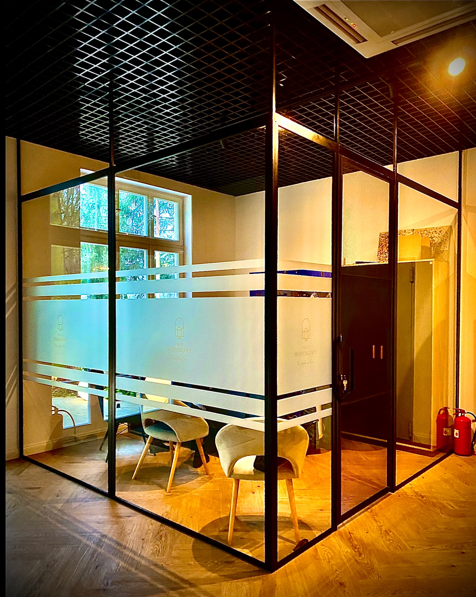 Nowoczesna, przeszklona konstrukcja biurowa z czarnymi profilami i matowymi pasami z logo, oświetlona ciepłym światłem, z widocznymi krzesłami wewnątrz.