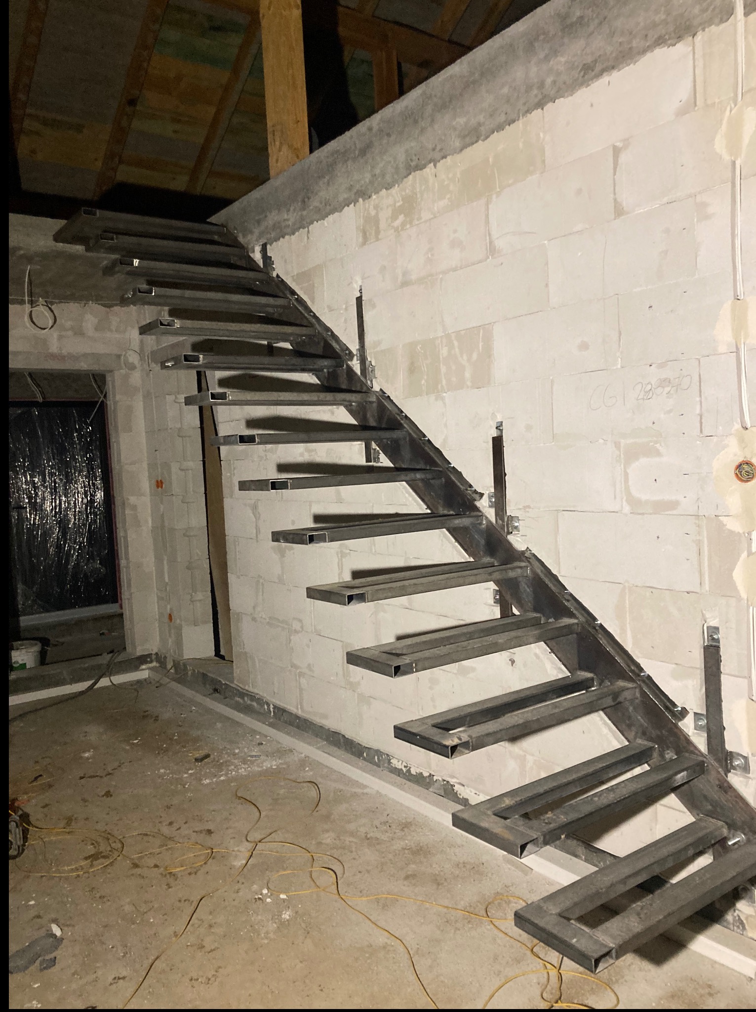 Surowe, metalowe schody z profili kwadratowych, w trakcie montażu w surowym wnętrzu z białych bloczków. Widoczne kable elektryczne na podłodze i przygotowane mocowania.