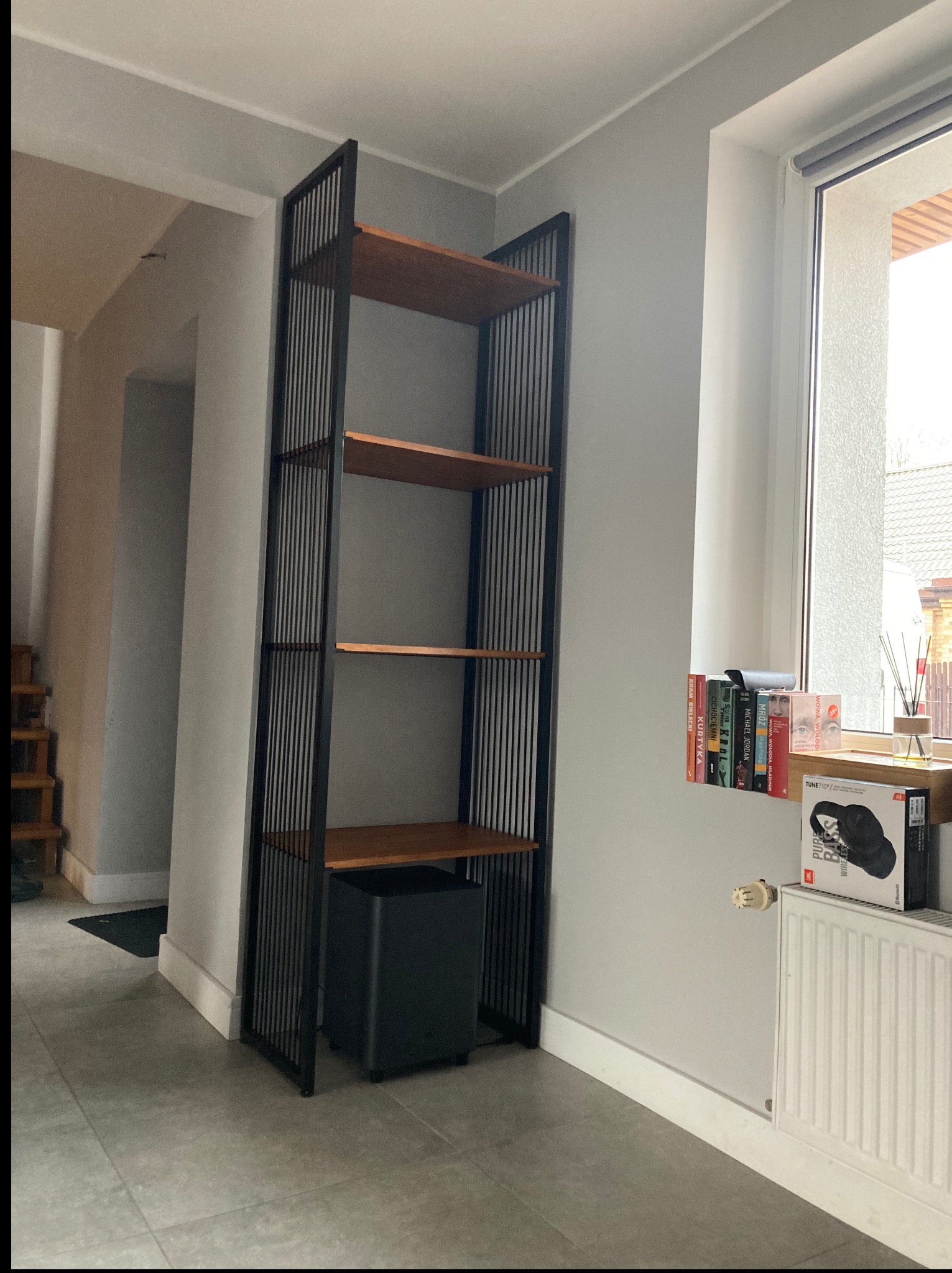 Regał metalowy z drewnianymi półkami w nowoczesnym wnętrzu. Czarna rama regału z pionowymi prętami. Na dolnej półce czarny subwoofer. Obok okno z książkami i grzejnik.